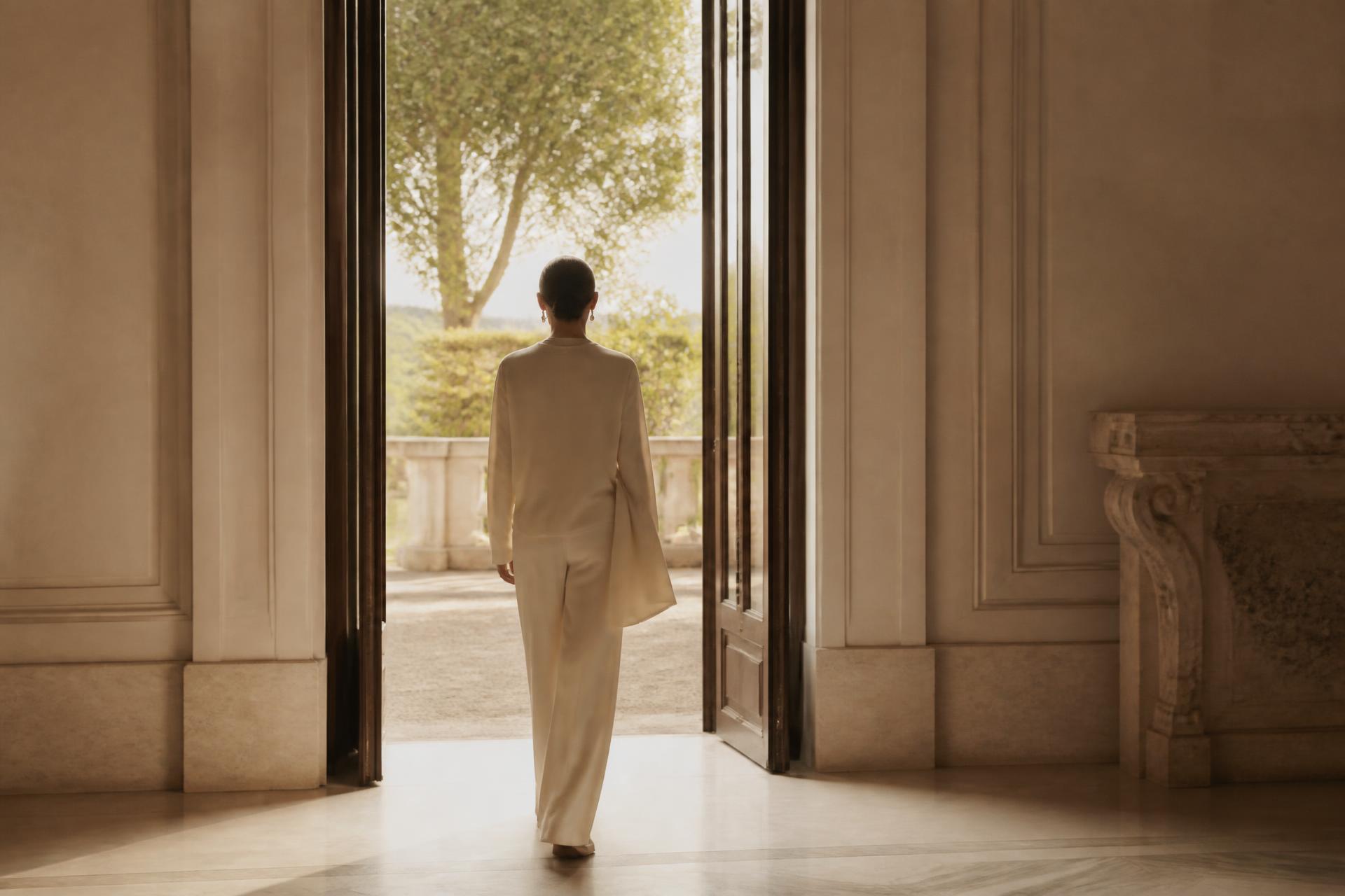 A woman in cream stepping through a tall open doorway into daylight, seen from behind