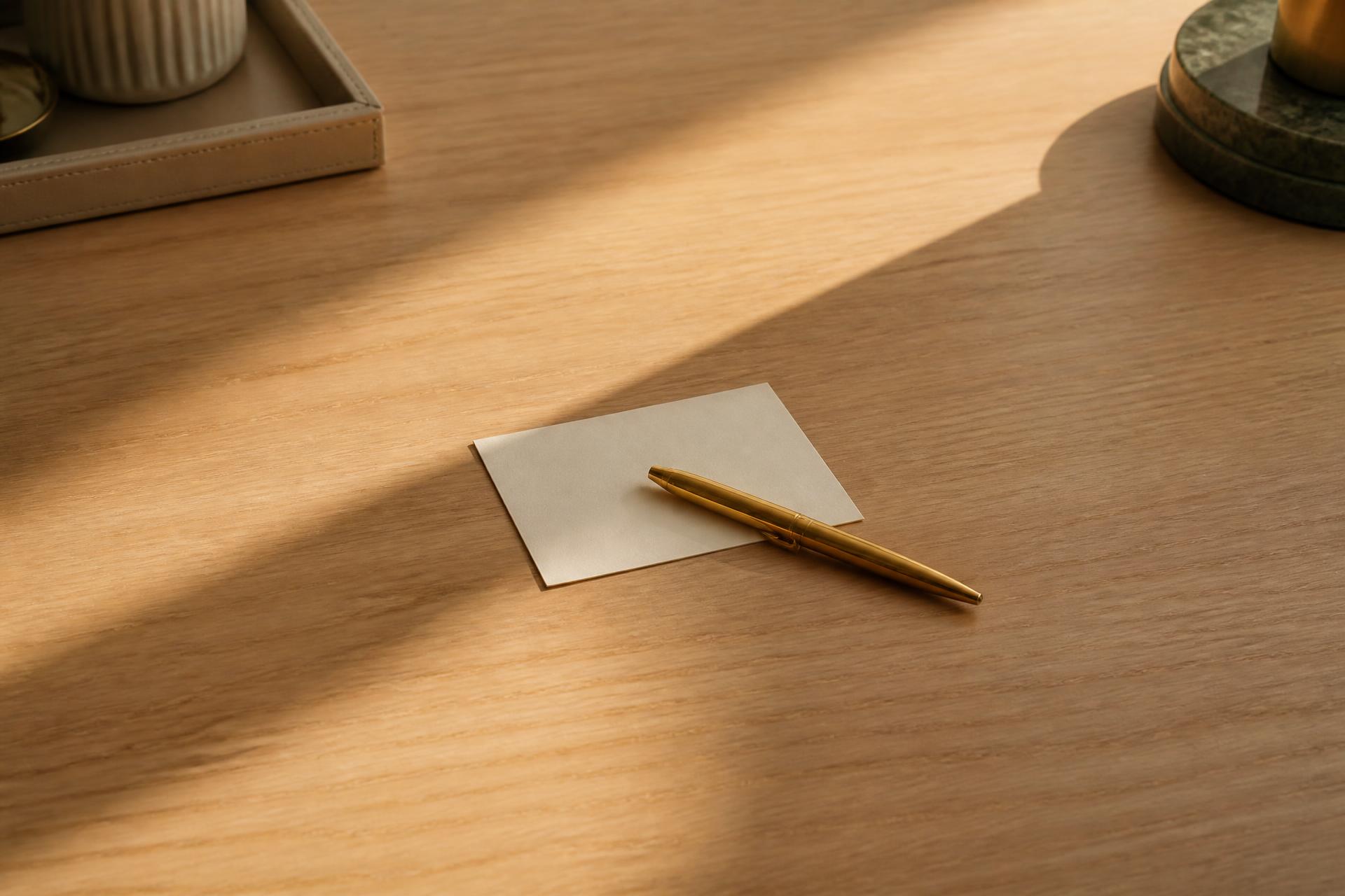 A cleared oak desk with a single blank cream card and one gold pen in warm morning light